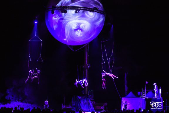 Des acrobates suspendus dans les airs sous une grande sphère lumineuse violette.
