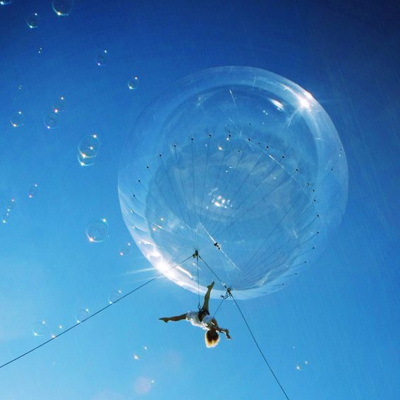 Une artistye suspendu à une bulle géante dans un ciel bleu parsemé de bulles.