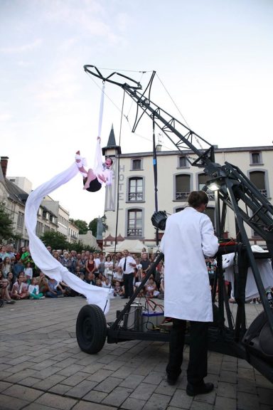 Un artiste suspendu, vêtu de blanc, performe avec une structure métallique sur une place.