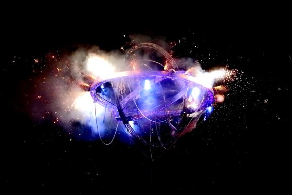 Un véhicule futuriste avec des lumières et des effets pyrotechniques sur fond noir.