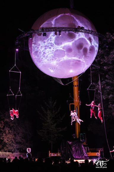 Des acrobates suspendus en balançoire devant un globe lumineux dans un spectacle nocturne.