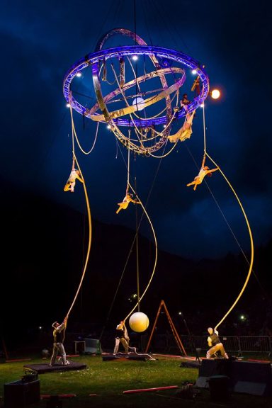 Performers manipulate a large, illuminated structure with suspended elements in a dark sky.