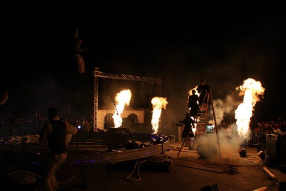 Scène nocturne avec flammes et effigies sur fond de festival ou d'événement.
