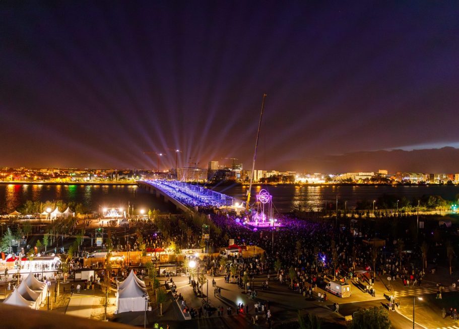 Vue nocturne d'une ville illuminée avec des rayons de lumière et un pont en arrière-plan.