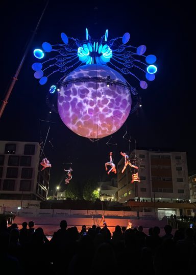 Une installation lumineuse avec des acrobates suspendus sous un globe flottant.