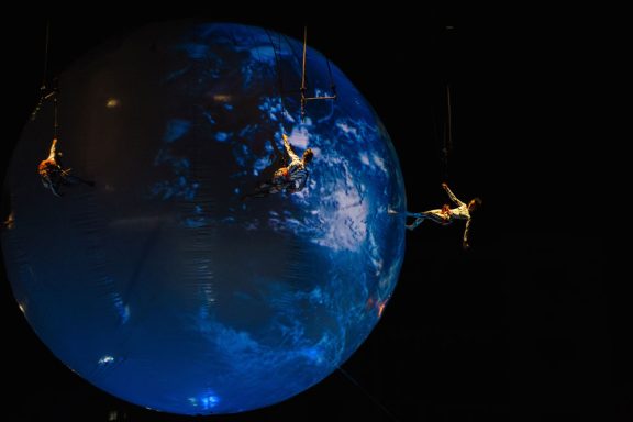 Performers are suspended near a glowing, stylized globe in a dark setting.