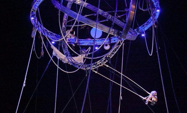 Un artiste de cirque suspendu à une structure en métal, entouré de lumière.