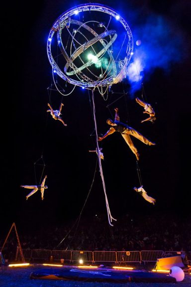 Artistes acrobatiques suspendus dans les airs, entourés de lumières vives.