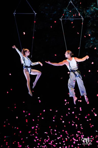 Deux artistes acrobates suspendus dans les airs, entourés de pétales et de lumières.