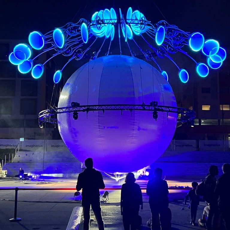 Installation artistique lumineuse avec un grand globe blanc et des éléments suspendus bleus.