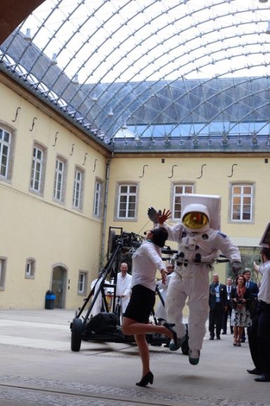 Une femme en jupe danse avec un astronaute dans un grand hall sous une verrière.
