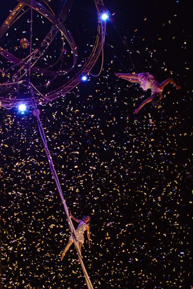 Des artistes acrobates se produisent sous un ciel étoilé, entourés de paillettes dorées.
