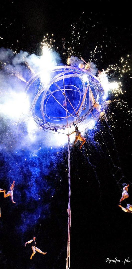 Artistes de cirque exécutant des acrobaties autour d'une structure illuminée par des feux d'artifice.