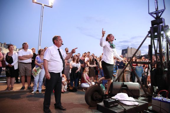 Une foule attentive assiste à un spectacle en plein air, avec un artiste sur scène.