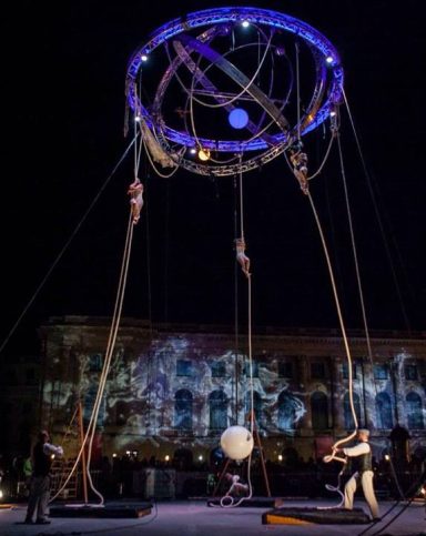 Performance artistique avec des acrobates, une structure circulaire et un globe au sol.
