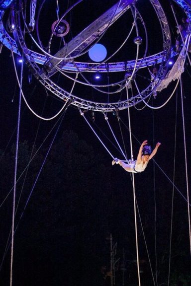 Artiste de cirque suspendu dans les airs, entouré de cercles métalliques et de câbles.