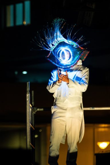 Personne en costume blanc tenant un symbole lumineux en forme d'œil sur scène.