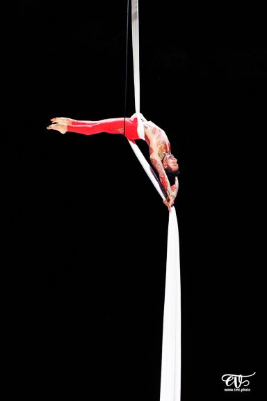 Artiste de cirque en apesanteur, suspendu à un tissu, en tenue rouge sur fond noir.