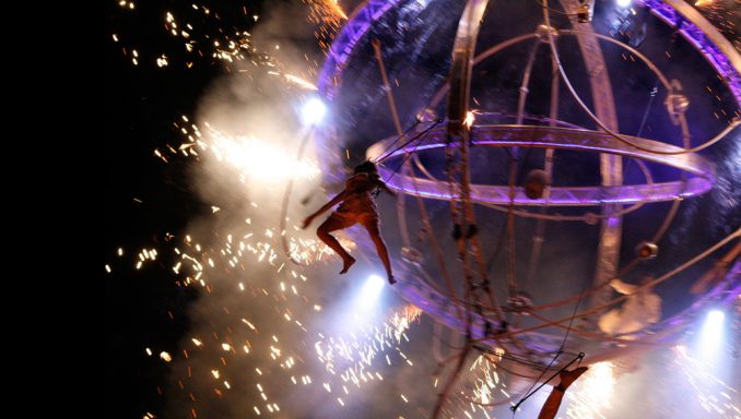 Artiste acrobatique suspendu dans une sphère, entouré d'étincelles et de lumière.