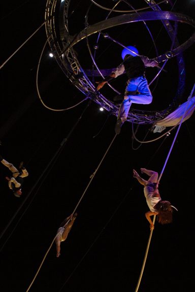 Artistes de cirque s'élevant dans les airs, suspendus par des cordes, sur fond sombre.