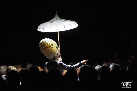Person en costume avec un grand chapeau et un parasol, se tenant devant une foule.