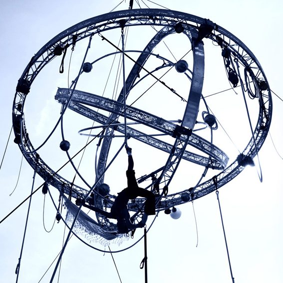 Artiste acrobatique suspendu dans une structure en forme de sphère, ciel en arrière-plan.