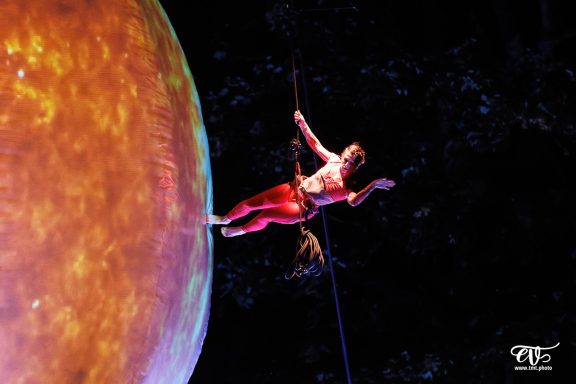 Artiste acrobatique suspendu, évoluant près d'une grande planète colorée.