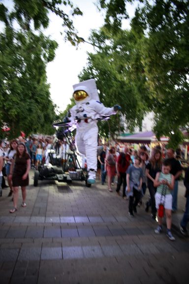 Un personnage géant en costume d'astronaute défile devant une foule sur une rue animée.