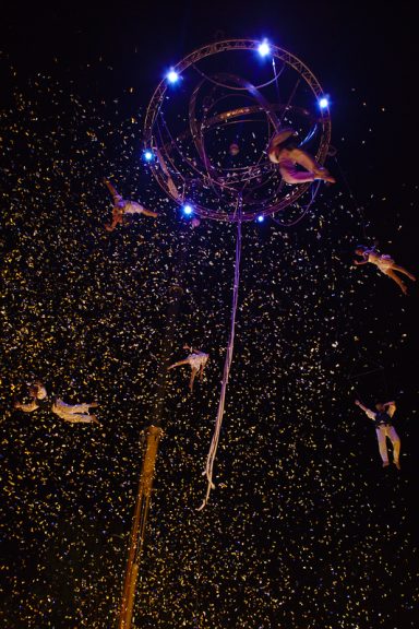 Six acrobates suspendus dans les airs, illuminés par des lumières, entourés de confettis.