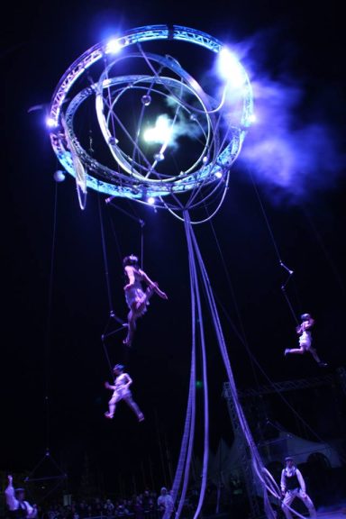 Acrobates suspendus dans les airs, illuminés par des lumières colorées.